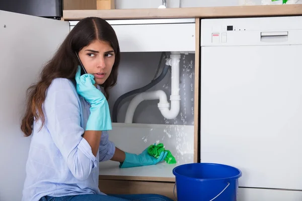 Femme stressée appelant un plombier d'urgence pour une fuite d'eau sous son évier.